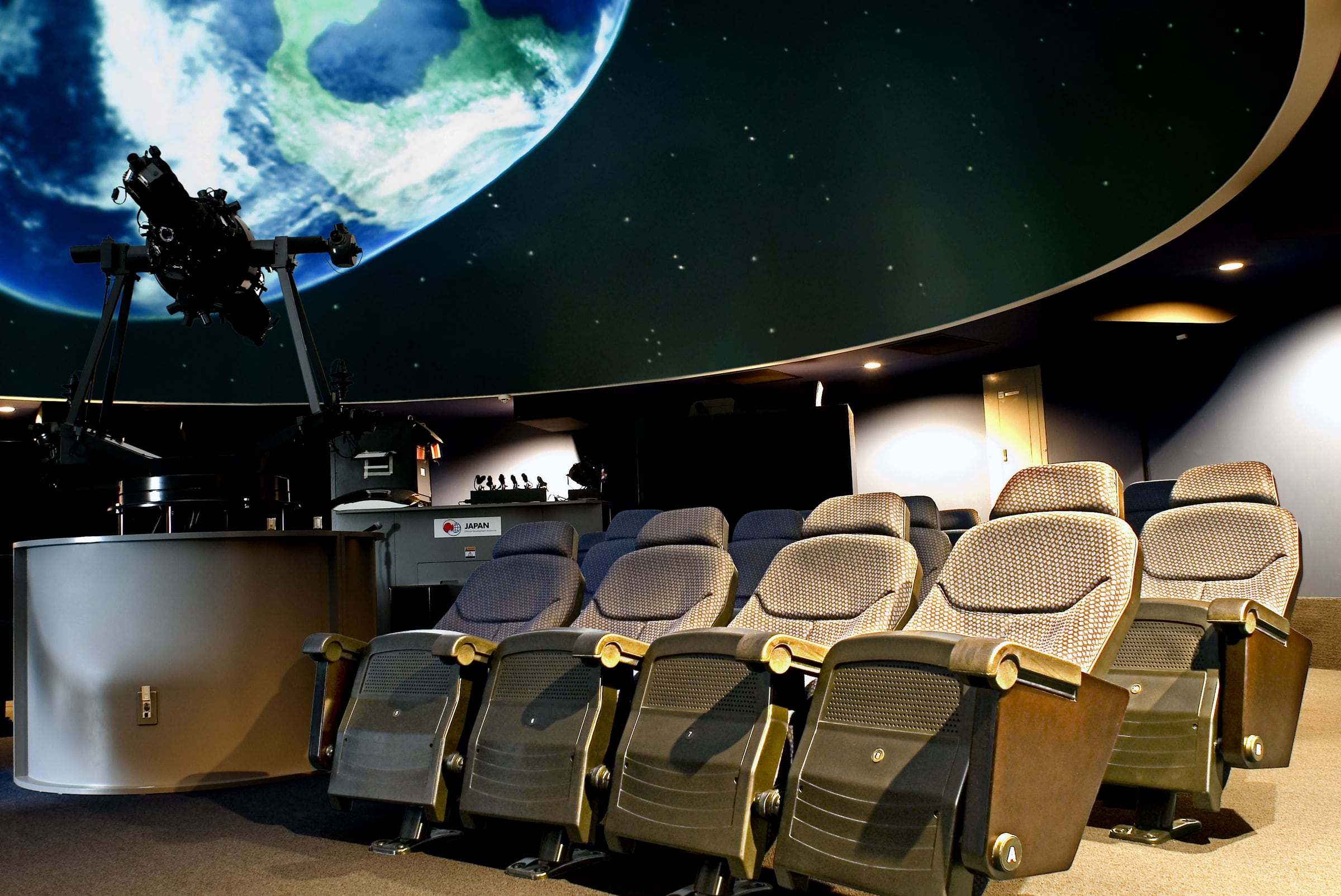 Butacas en primer plano en la sala del Planetario de la UCR, con el proyector y la proyección de la Tierra en la cúpula