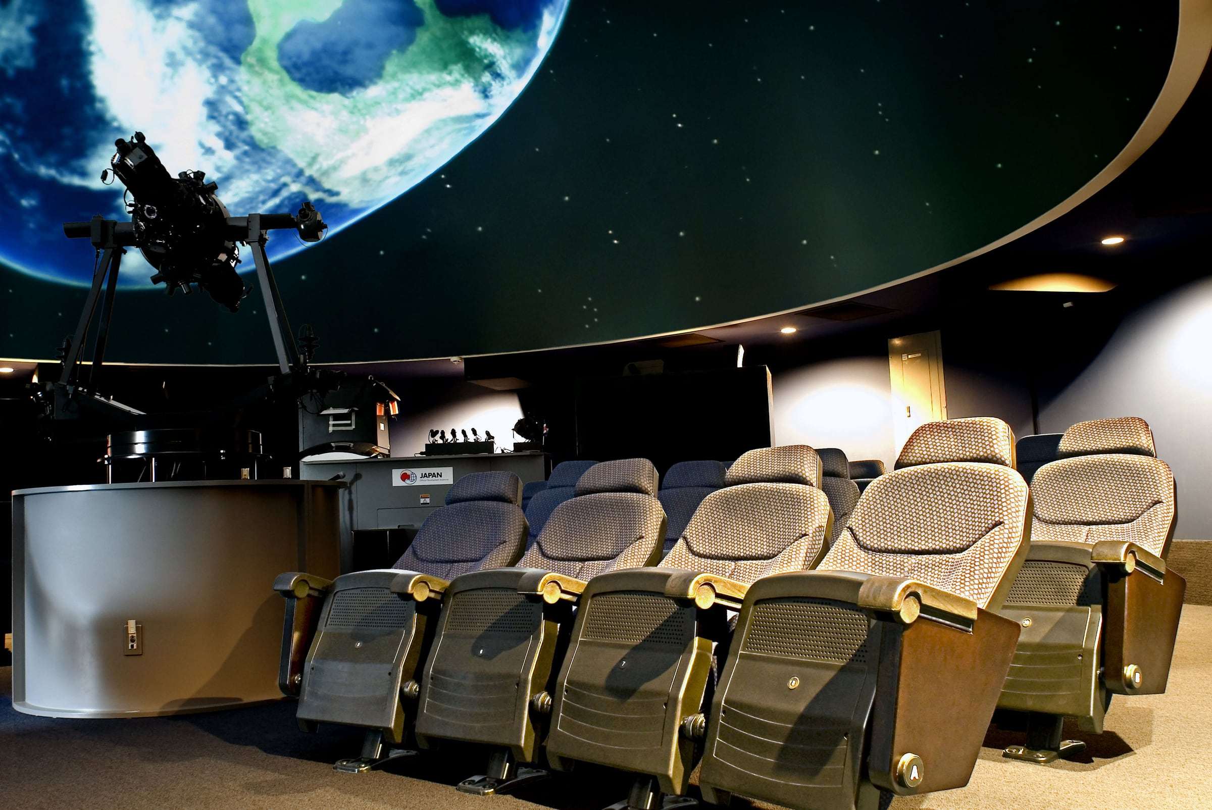 Butacas en primer plano en la sala del Planetario de la UCR, con el proyector y la proyección de la Tierra en la cúpula
