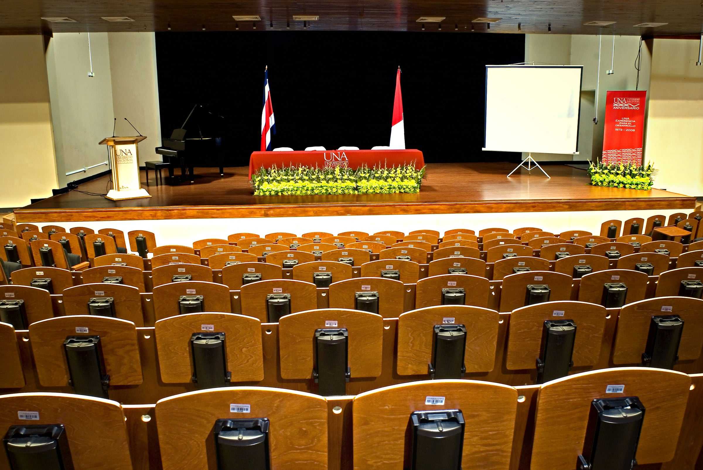 Butacas de madera frente al escenario del auditorio de la Universidad Nacional, con las banderas al fondo