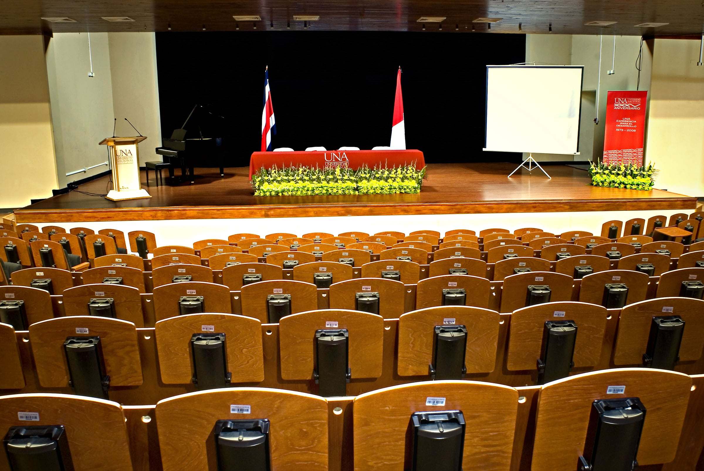 Butacas de madera frente al escenario del auditorio de la Universidad Nacional, con las banderas al fondo